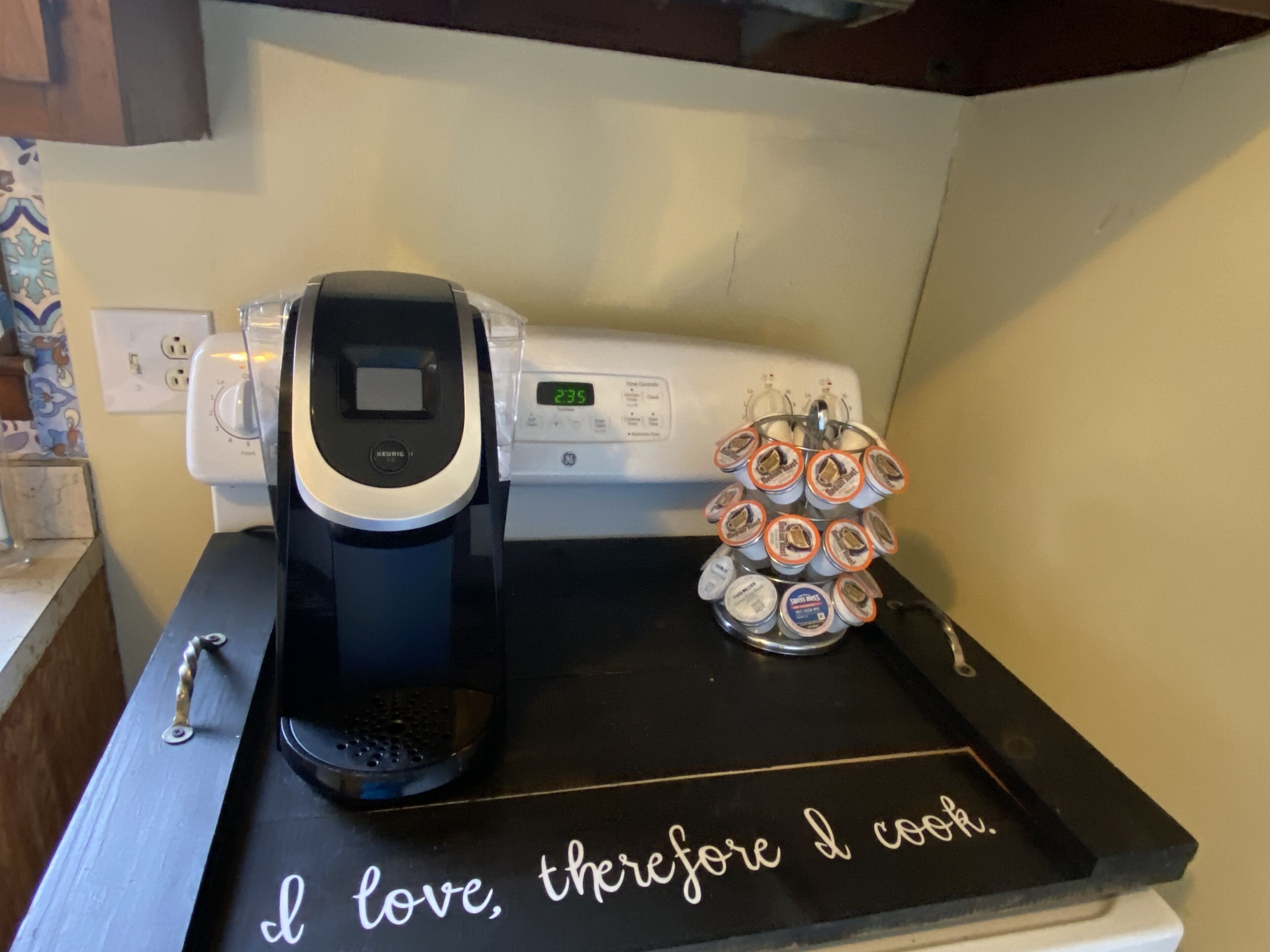 A kitchen counter with a coffee maker, a stack of coffee pods, and a sign that says "I love, therefore I cook."