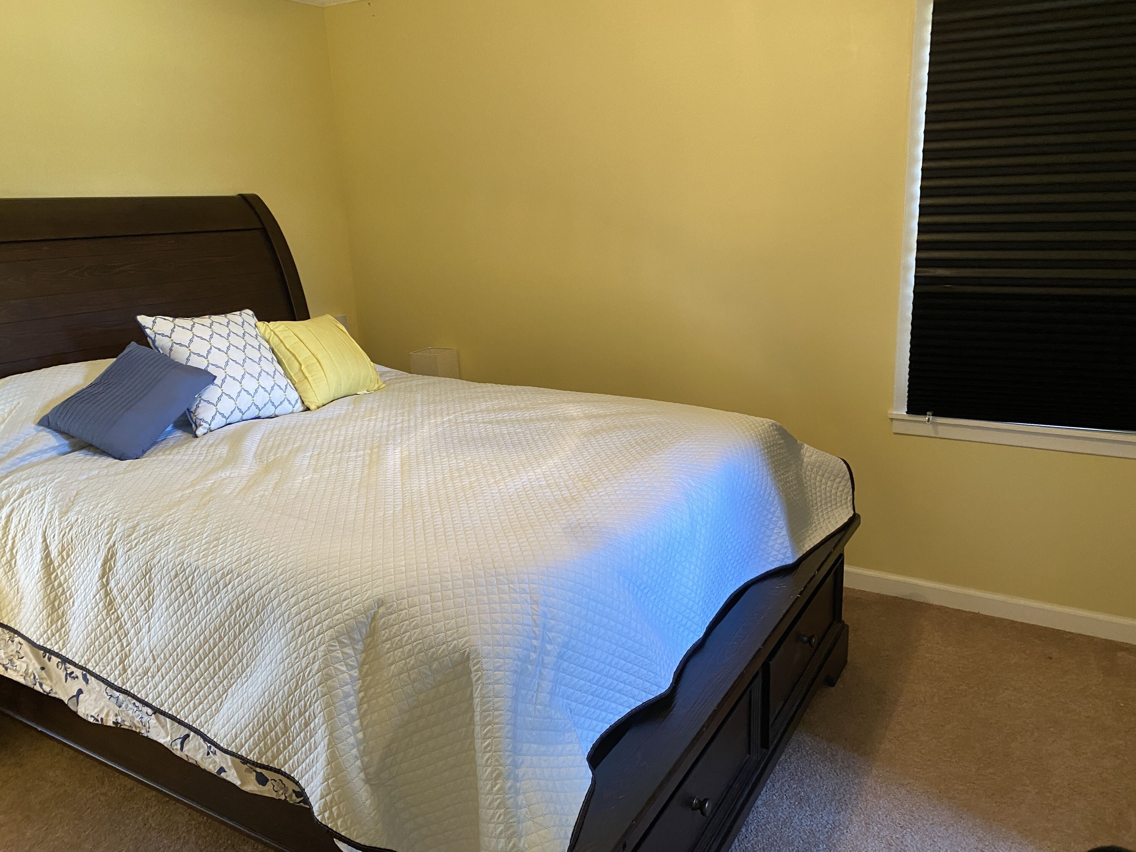 Een slaapkamer met een bed, kussens, een hoofdbord, een raam met zonwering en een vloerbedekking.