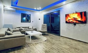 A modern living room with a sectional sofa, a coffee table, a TV mounted on the wall, and a sliding glass door leading to a balcony.