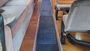 Interior de una camper con un pasillo central, asientos acolchados a ambos lados, una mesita y una zona de cocina en la parte trasera.