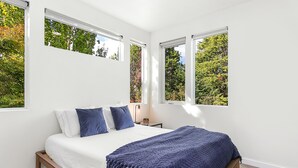 A bedroom with a bed, white walls, and large windows with a view of trees.