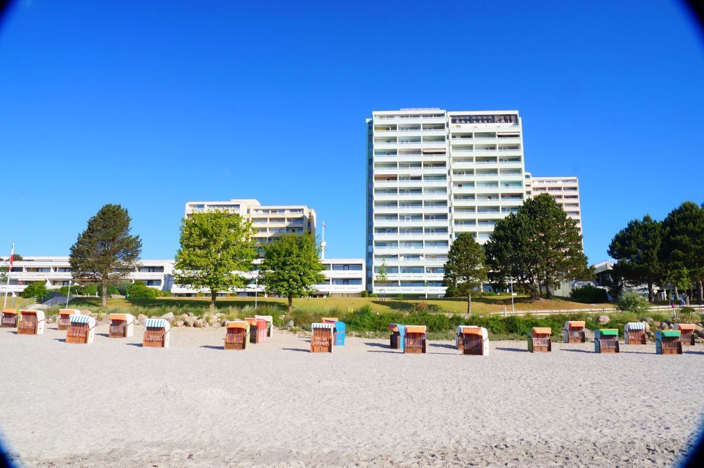 Un hotel frente a la playa con varias construcciones, zonas de estar al aire libre y un cielo azul despejado.