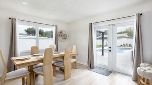 Een eethoek met een houten tafel en stoelen, een rond wandplankje en uitzicht op een zwembad door glazen deuren.
