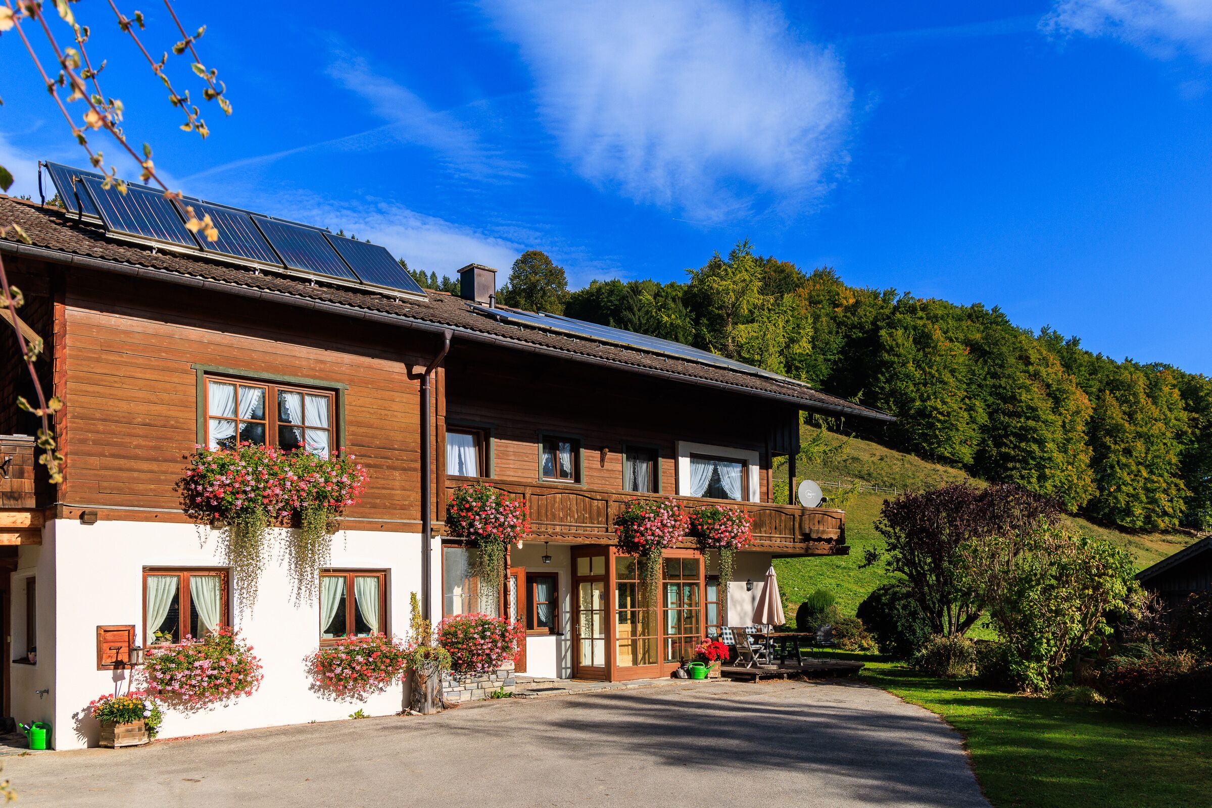 A two-story wooden building with large windows, a balcony with hanging flower baskets, and a paved driveway leading to a car.