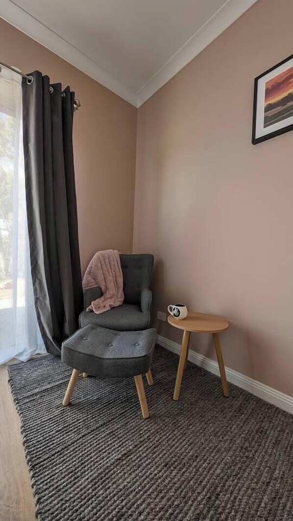 Une pièce d’angle avec un fauteuil gris, une petite table d’appoint en bois, un tapis gris et une fenêtre avec des rideaux foncés.