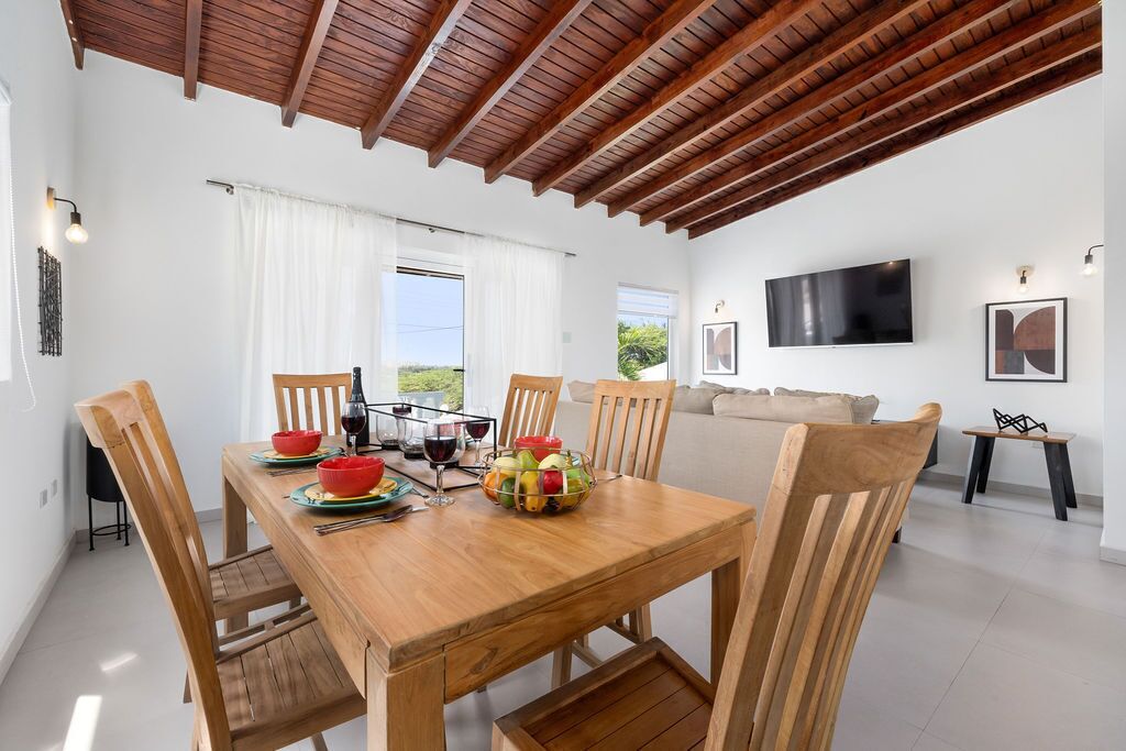 A dining area with a wooden table, chairs, and a fruit bowl. There is a sofa and a TV in the background.