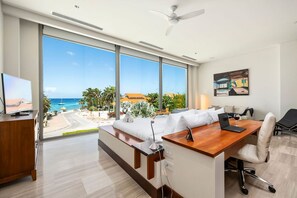 A hotel room with a large window offering a view of the beach and ocean, a wooden desk with a laptop, a bed with white linens, and a ceiling fan.