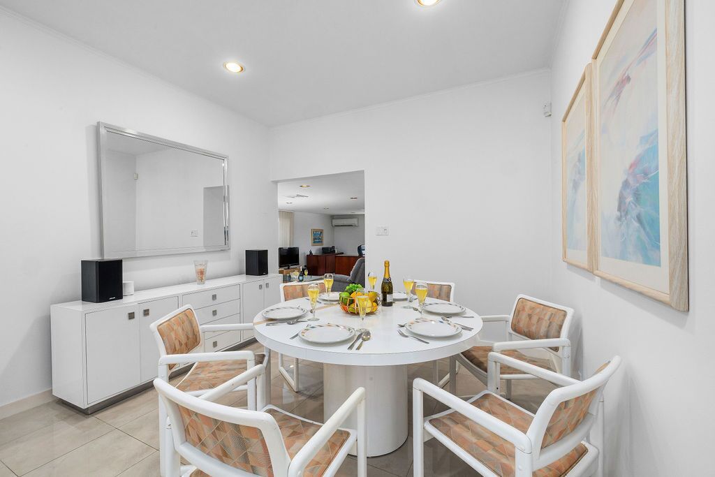 A modern dining room with a round table set for six, surrounded by white chairs with patterned cushions. A large mirror and framed artwork adorn the walls.