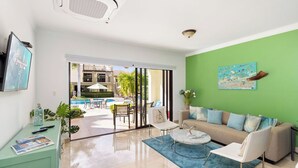 A modern living room with a green wall, a sofa, a coffee table, and a view of an outdoor pool area. - Ground Floor 1BR Condo w/ Pool & Resort Amenities (Noord)
