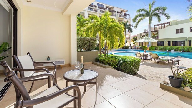 A balcony with a table, chairs, and a bottle of champagne. There is a pool with lounge chairs and palm trees in the background. - Ground Floor 1BR Condo w/ Pool & Resort Amenities (Noord)