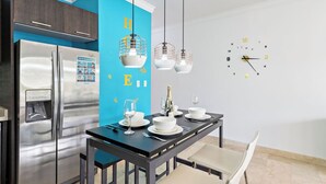 A modern kitchen with a blue accent wall, stainless steel appliances, and a dining area set for four with a clock on the wall. - Ground Floor 1BR Condo w/ Pool & Resort Amenities (Noord)