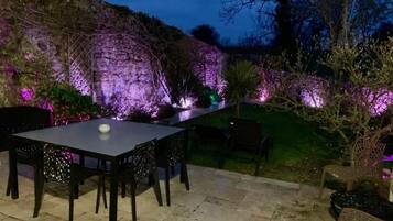 Une terrasse extérieure avec une table et des chaises, entourée de murs en pierre et agrémentée de plantes illuminées.