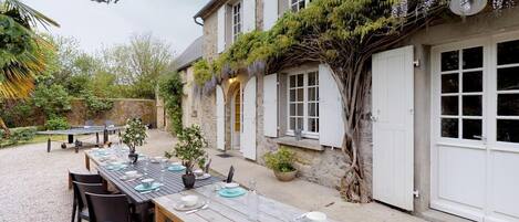 Un edificio in pietra con un'area pranzo all'aperto coperta, un grande albero e un cortile in ghiaia.