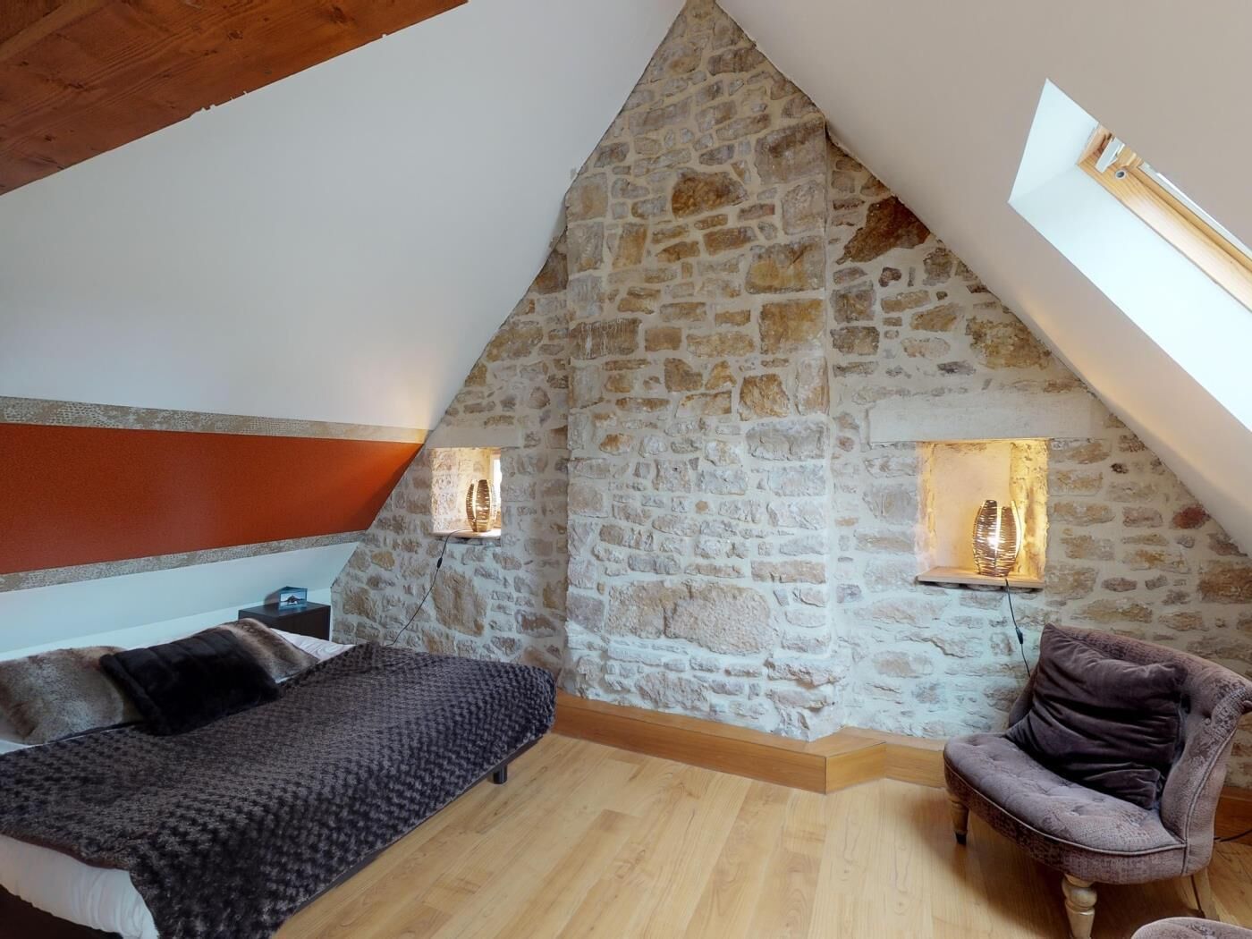A bedroom with a sloped stone wall, a bed, a chair, and two wall-mounted lights.