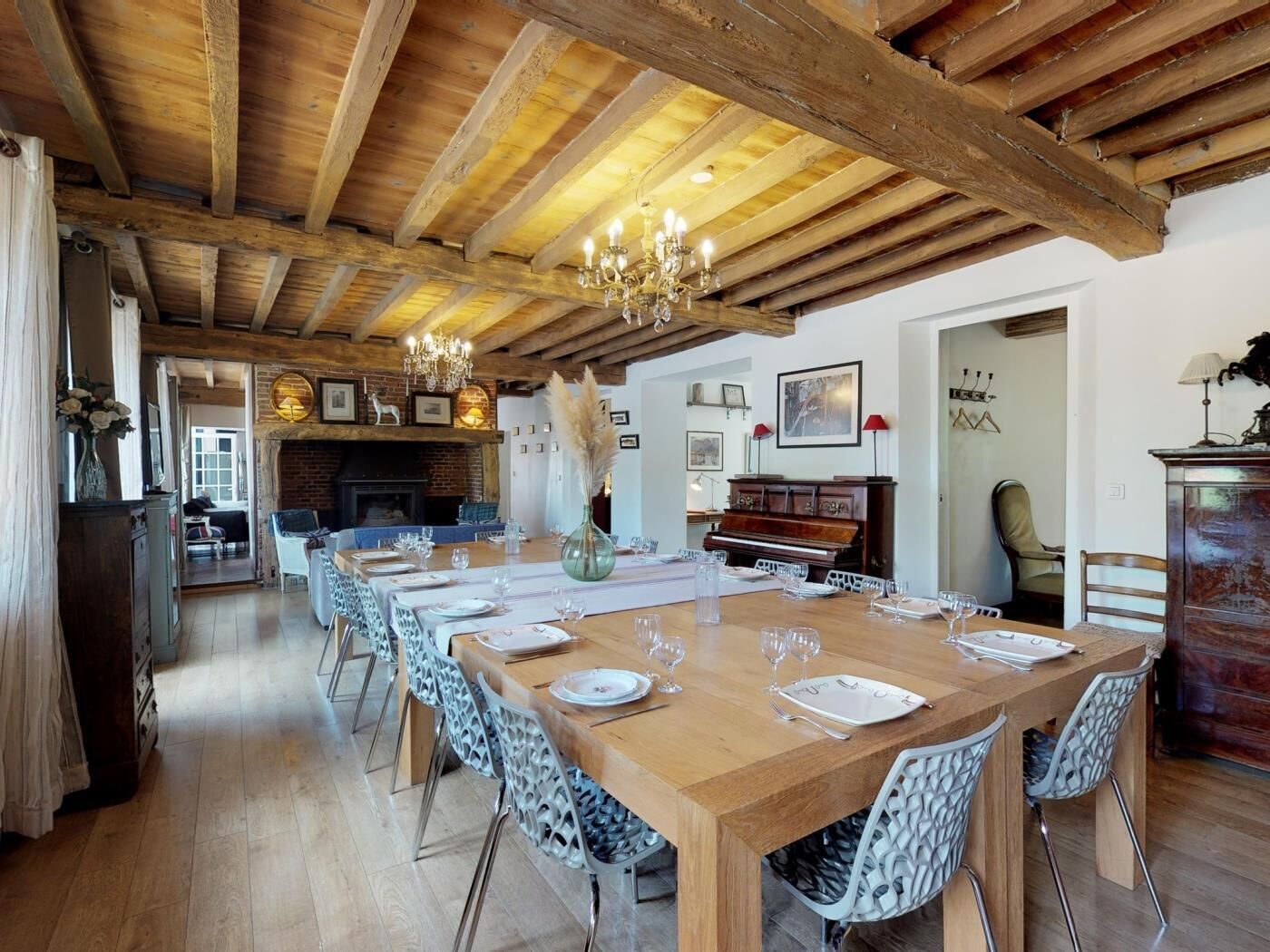 Un coin repas avec une longue table en bois dressée pour six personnes, entourée de chaises à motifs, d’un lustre et d’un piano en arrière-plan.