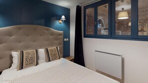 Un dormitorio con cabecera capitonada, cama con ropa de cama estampada, lámpara de pared y ventana con vista a una edificación de ladrillo.