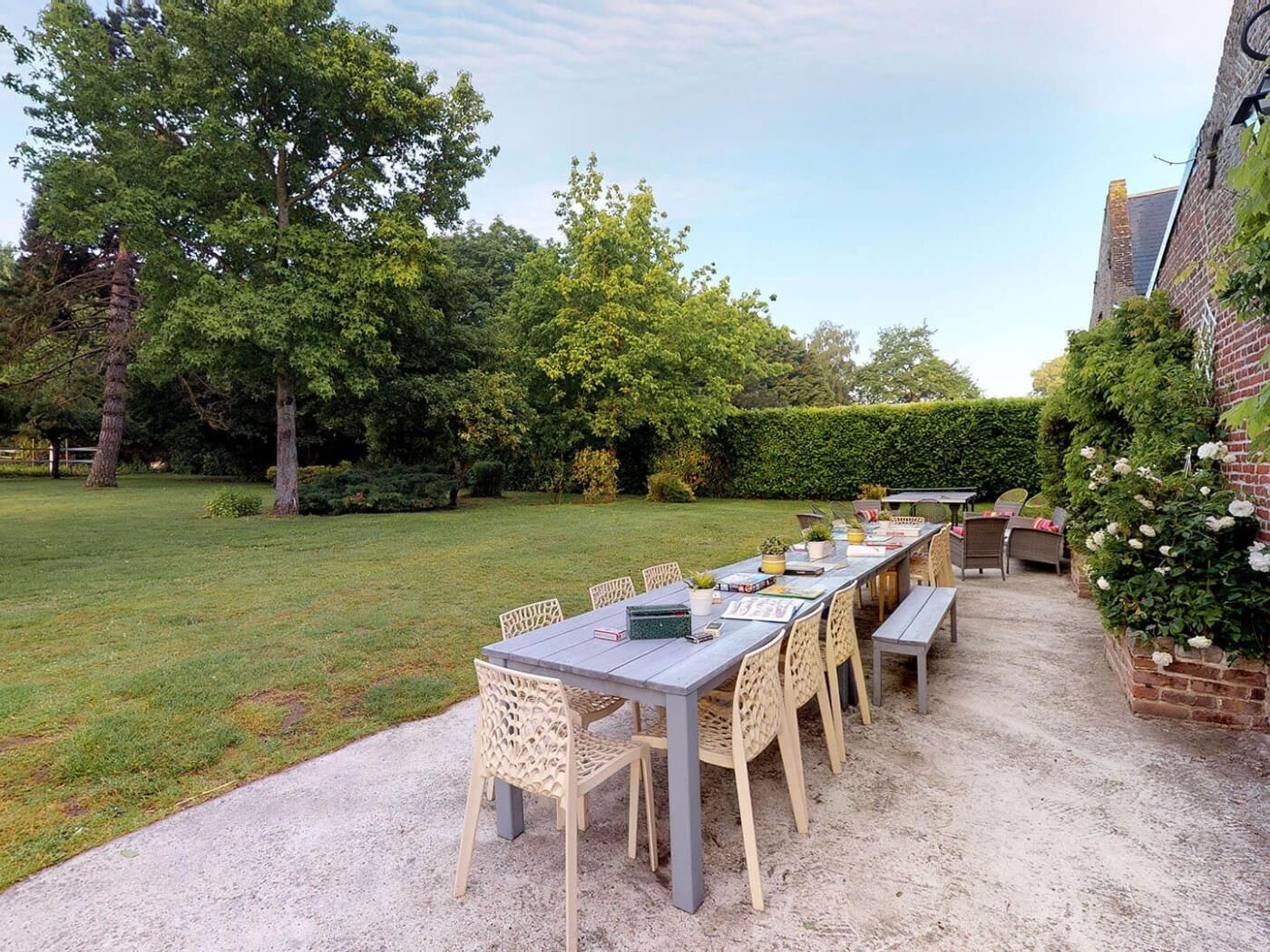An outdoor patio with a long table and chairs, surrounded by greenery and a brick wall.