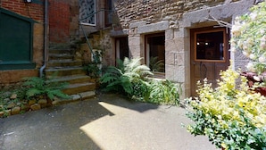 A stone building with a wooden door, a staircase, and a small garden area with ferns and flowering plants. - Cour du paradis 8 pers : billiards, baby, 30 min sea (Villedieu-les-Poêles)