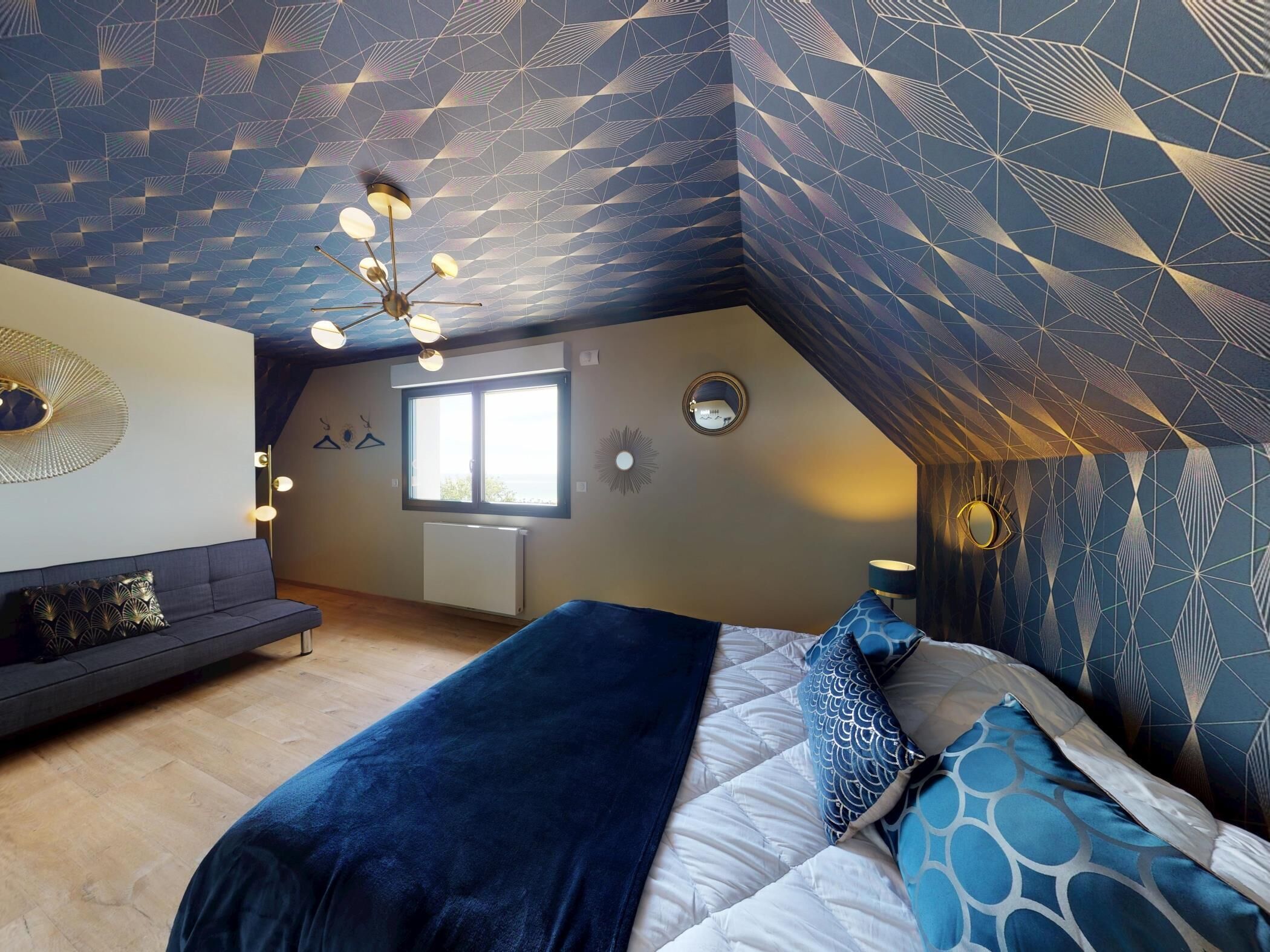 Une chambre avec un plafond en pente, un lit agrémenté d’oreillers à motifs, un canapé et une fenêtre.