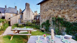 A stone building with a thatched roof, a wooden table set for a meal, and a garden area with a fence and greenery. - Villa Paradis 14 people : waterfront garden (Villedieu-les-Poêles)