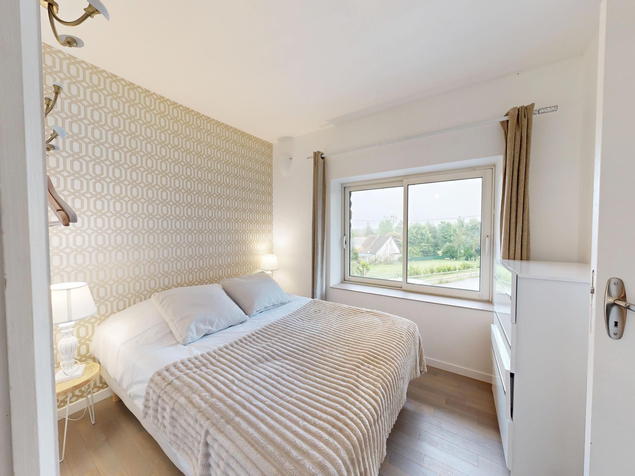 A bedroom with a bed, a bedside table, a lamp, a window with curtains, and a view of the outside.