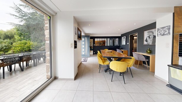 Een moderne eethoek met een grote tafel en gele stoelen, een keuken met een donkere achterwand, en uitzicht op groen buiten.