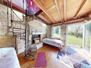 Une chambre à coucher rustique avec une cheminée en pierre, un escalier en bois, un lit et une grande fenêtre donnant sur l’extérieur.