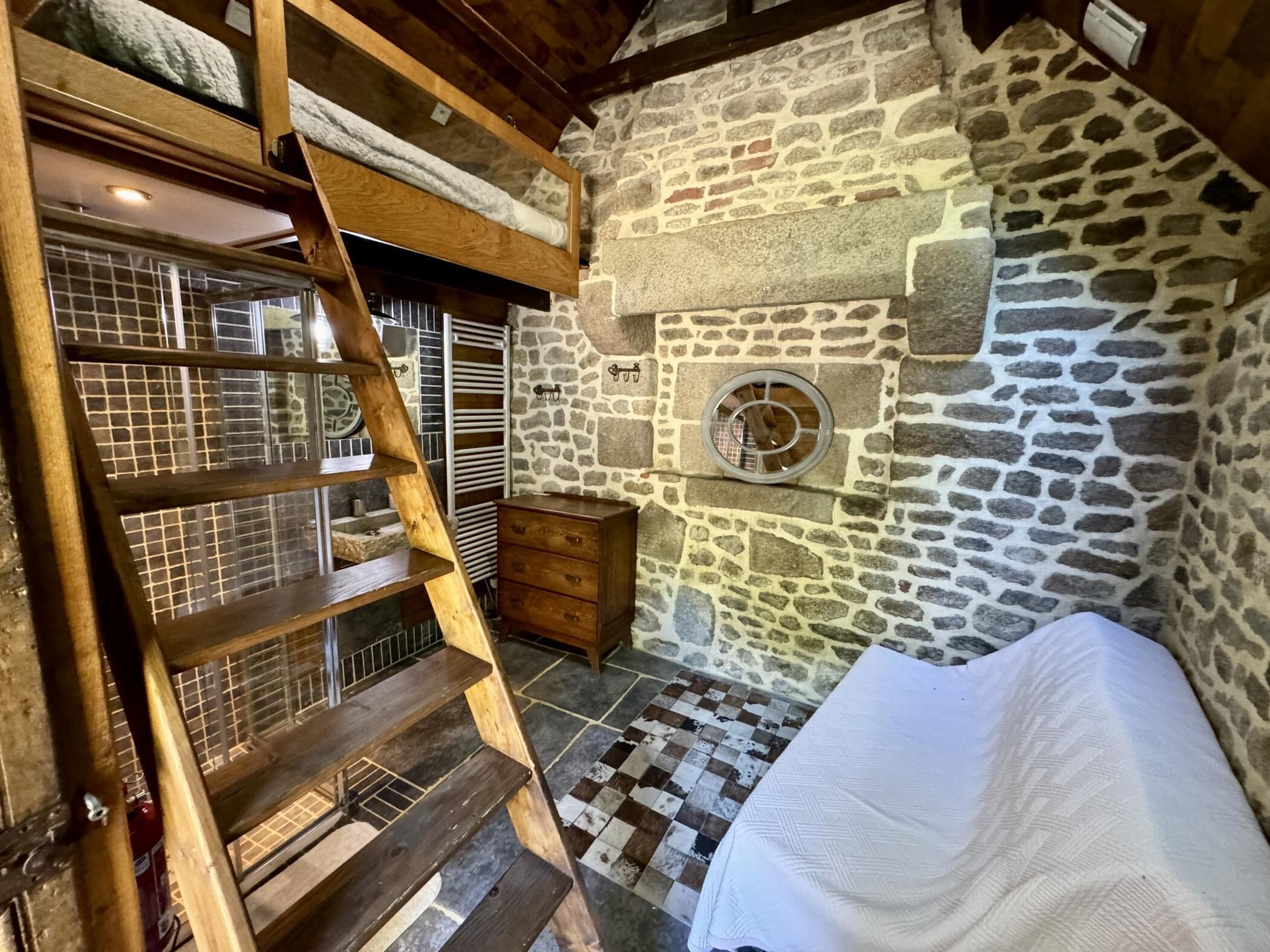 A rustic room with a stone wall, a wooden ladder, a bed, and a small wooden dresser.
