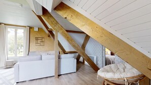 Dormitorio tipo loft con techo de madera, un sofá blanco, una silla de mimbre y vistas al exterior.