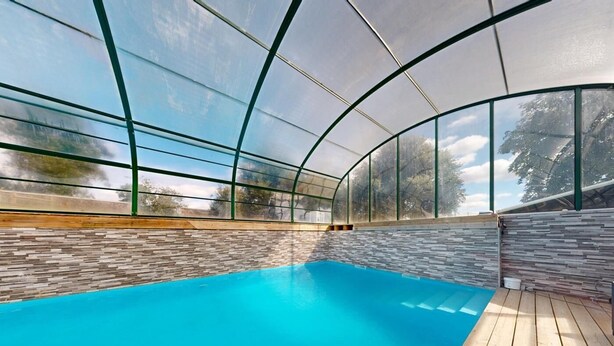 Una piscina interior con techo de vidrio transparente, cubierta de madera y pared de piedra.