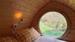 Un dormitori acollidor amb un sostre de fusta, un finestre rodó amb vista a vegetació, un llit amb roba de llit a quadres i una làmpada penjada a la paret.