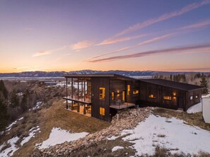 Eine moderne Holz-Blockhütte mit einer gläsernen Veranda, umgeben von verschneiter Landschaft und Bergen im Hintergrund.