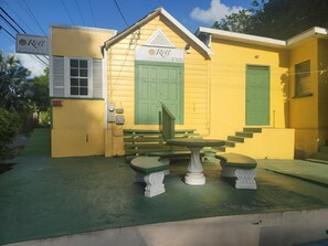 A yellow building with a green door and window shutters, a bench, and a table in front.
