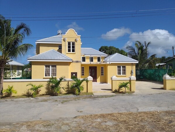 A two-story yellow building with a central entrance, surrounded by small palm trees and a paved area in front.