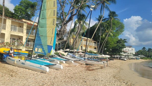 Une plage avec de nombreux voiliers et des parasols, des palmiers et un bâtiment doté de balcons.