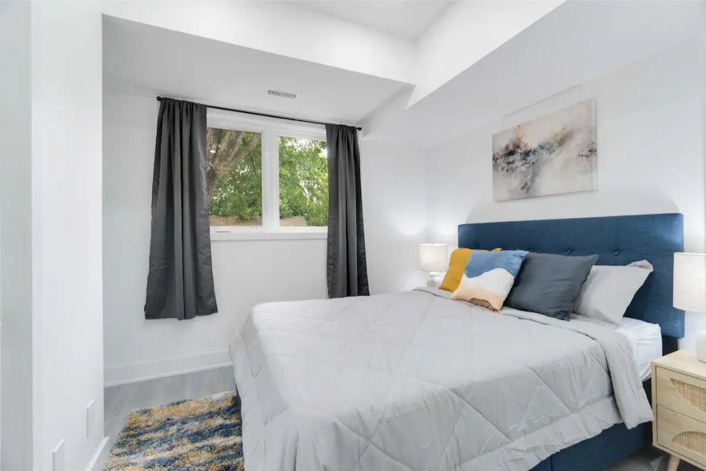 Une chambre à coucher avec un lit, une fenêtre avec des rideaux, une table de chevet avec une lampe et un tableau abstrait accroché au mur.