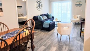 A living room with a sofa, a dining table with chairs, a ceiling fan, and a wall clock.