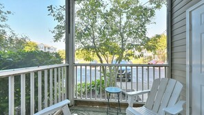 A porch with two white Adirondack chairs, a small round table, and a view of a street with cars and trees.