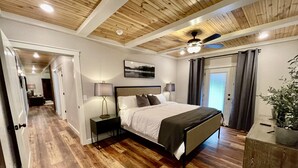 A bedroom with a wooden ceiling, a bed with a headboard, bedside lamps, a nightstand, a potted plant, and a view of a hallway. - Mountainside Luxury Cottage 2—LOCATION! LOCATION! (Mena)