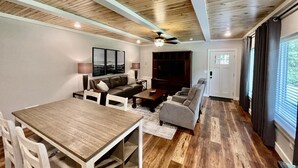 A living room with a wooden ceiling, a large TV, a sofa, a dining table, and a window with curtains. - Mountainside Luxury Cottage 2—LOCATION! LOCATION! (Mena)