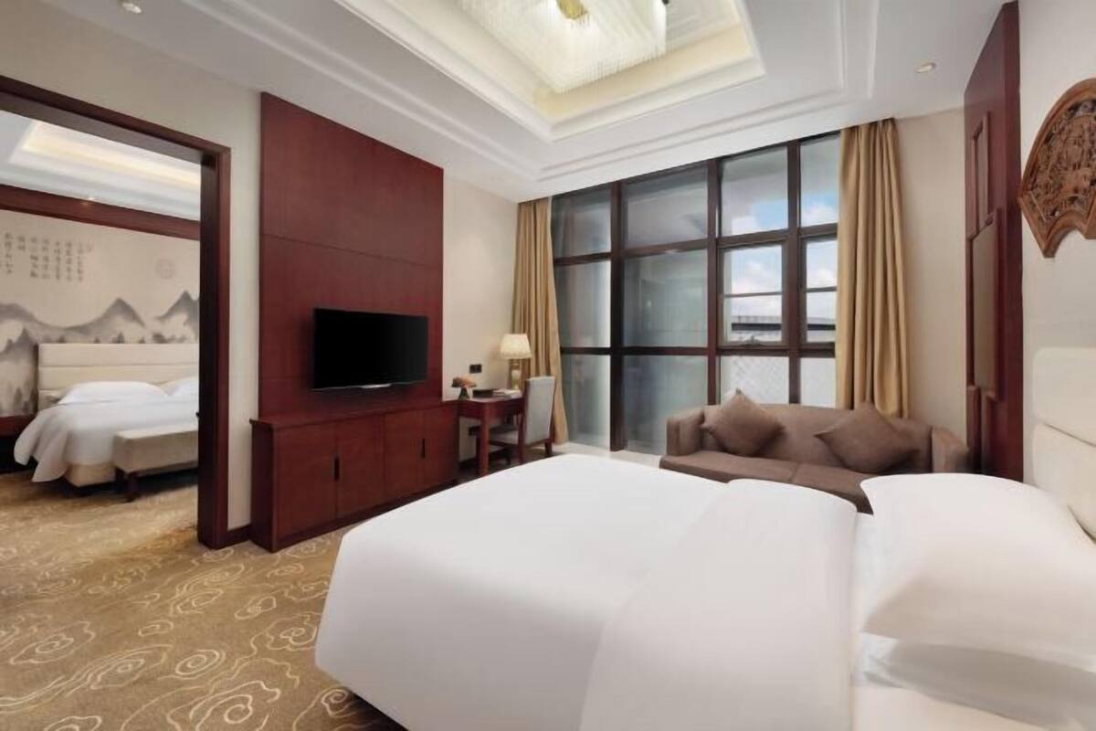 A hotel room with a large bed, a television, a desk with a lamp, and a view of the outdoors through large windows.