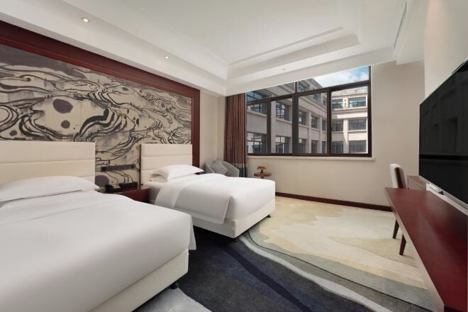 A hotel room with two beds, a large window, a flat-screen TV, and a marble-patterned wall.