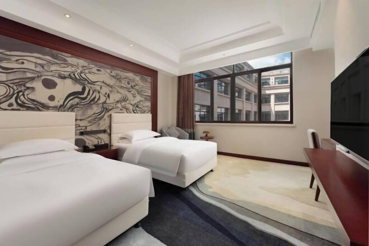 A hotel room with two beds, a large window, a flat-screen TV, and a marble-patterned wall.