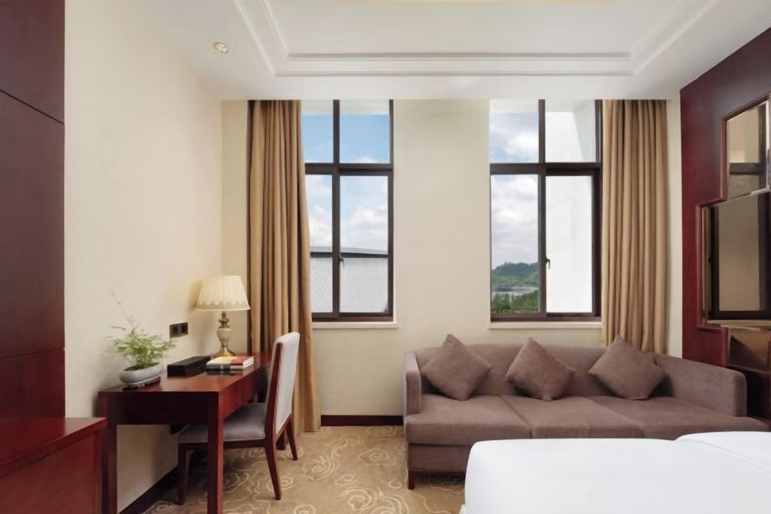 A hotel room with a bed, a desk with a lamp, a chair, a couch, and two windows with curtains.