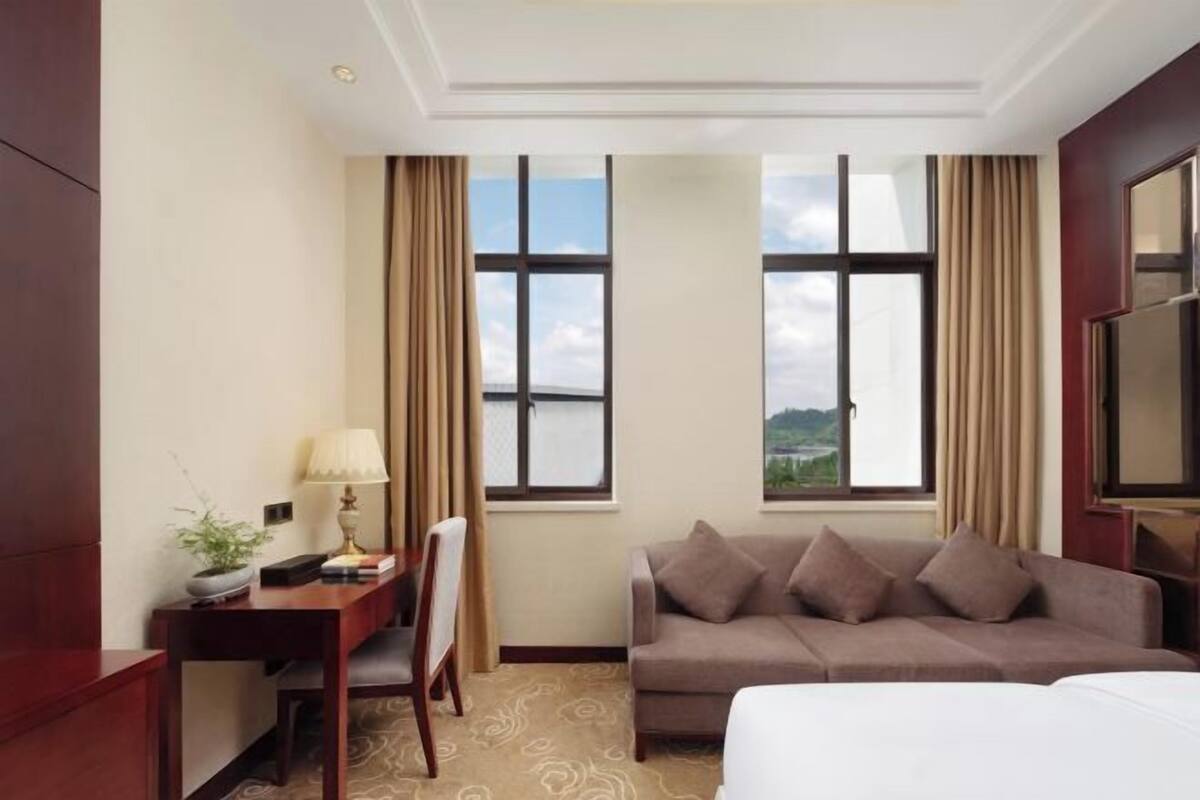 A hotel room with a bed, a desk with a lamp, a chair, a couch, and two windows with curtains.