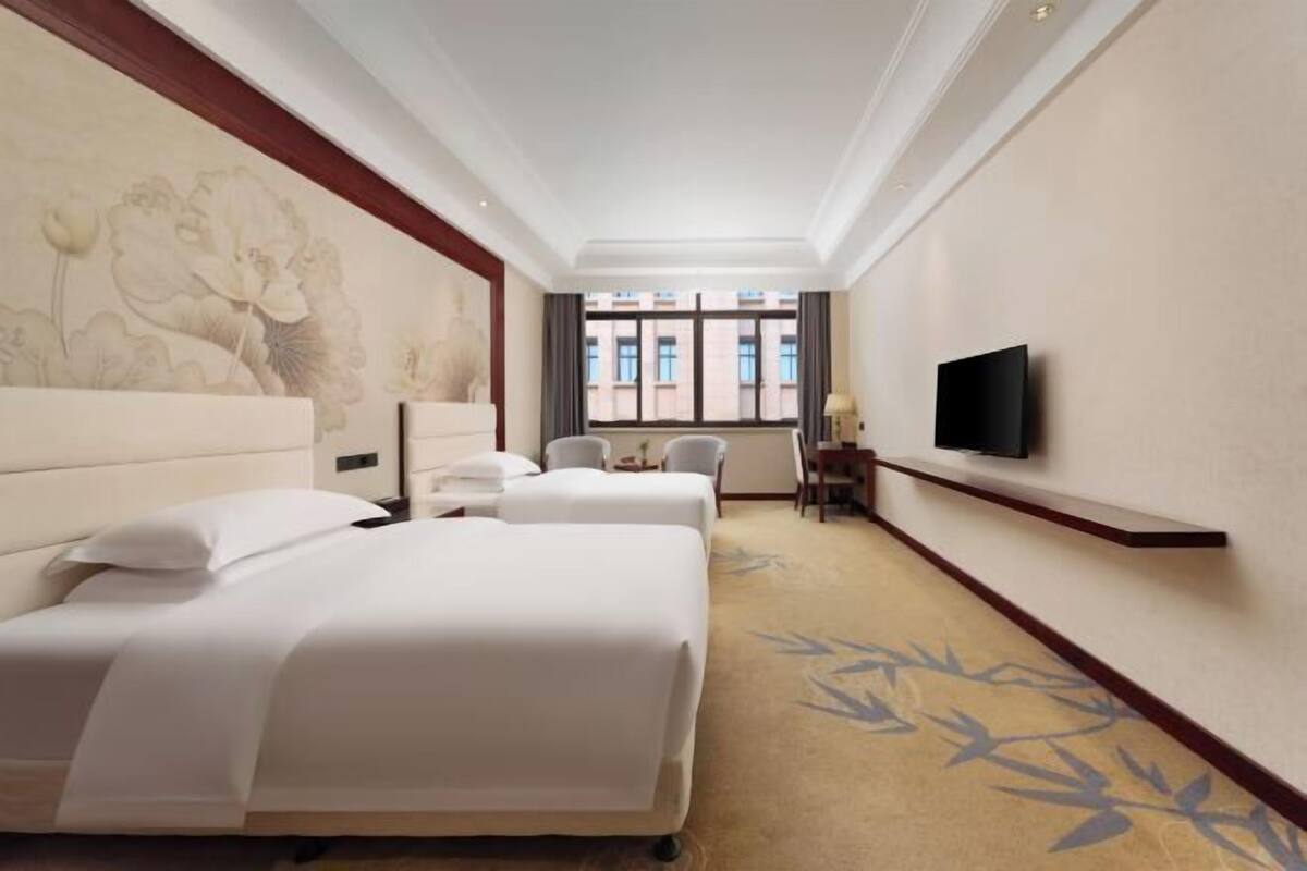 A hotel room with two beds, a television on the wall, a desk with a chair, and a window with a view of buildings.