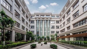 Un bâtiment moderne doté d’une façade centrale en verre, entouré d’un aménagement paysager symétrique et comprenant plusieurs étages.