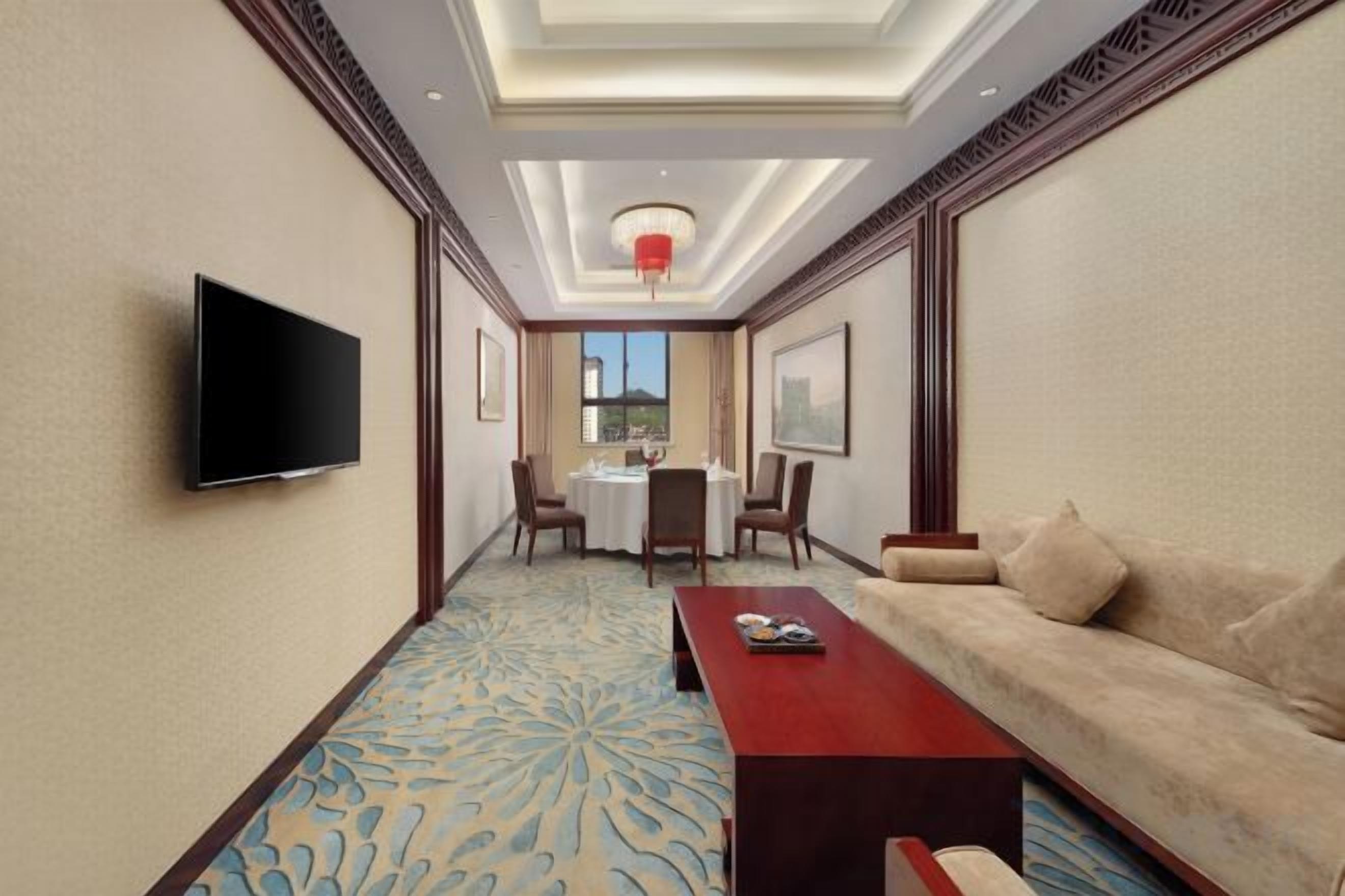 Ein Hotelzimmer mit Fernseher, Sofa, Couchtisch, Esstisch mit Stühlen und einem gemusterten Teppich.