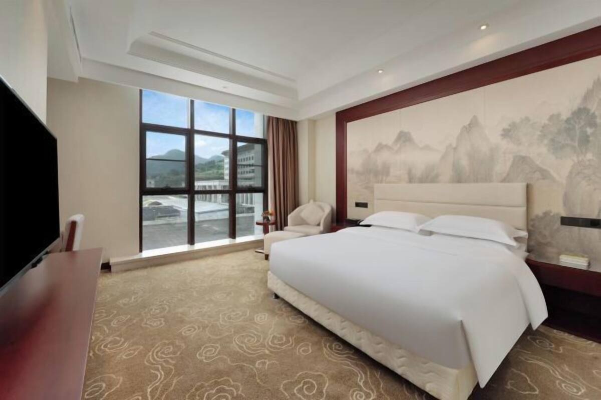A hotel room with a large bed, a desk, a chair, a television, and a large window with a view of the outside.