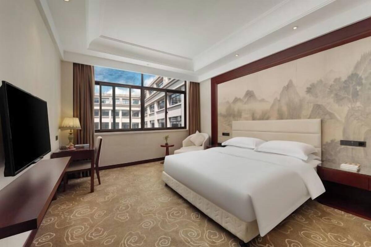 A hotel room with a large bed, a television, a desk with a lamp, a chair, and a window with a view of buildings.